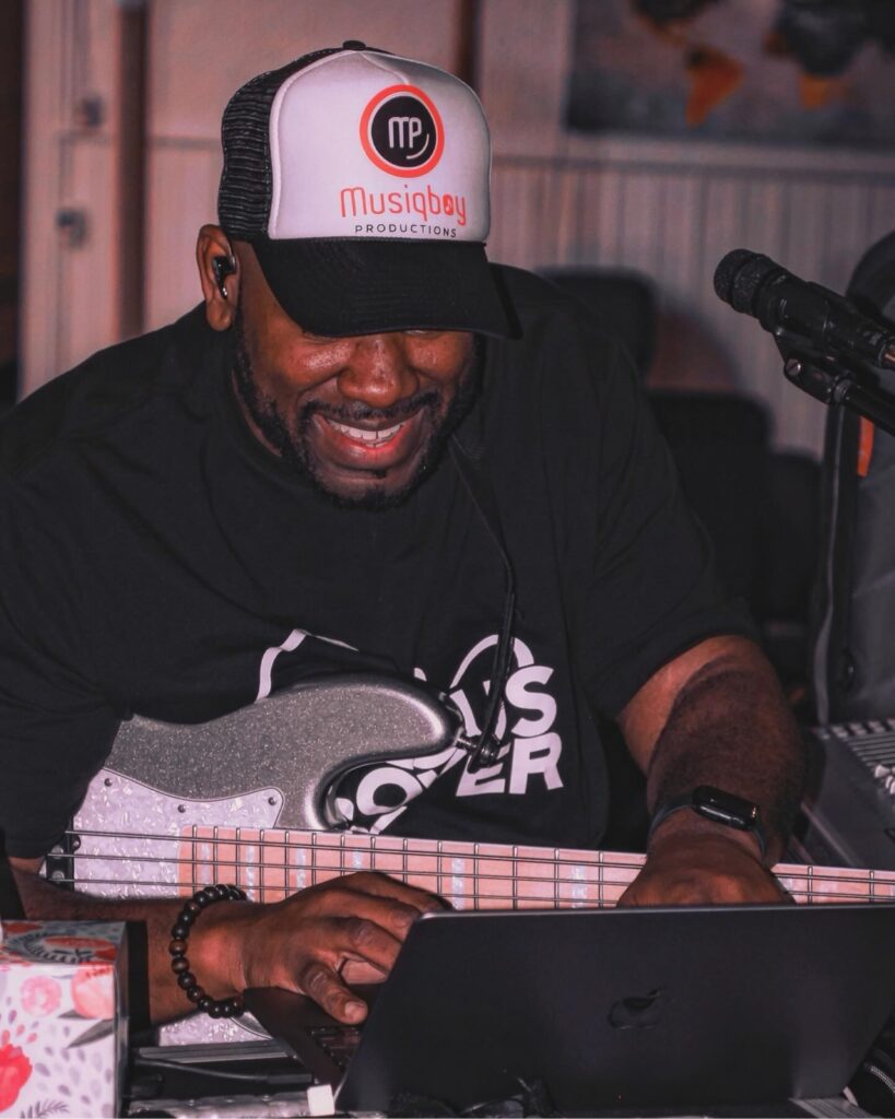 Jay Bratten wearing a Musiqboy Productions trucker cap, smiling while working on a laptop with a silver sparkle bass guitar in hand