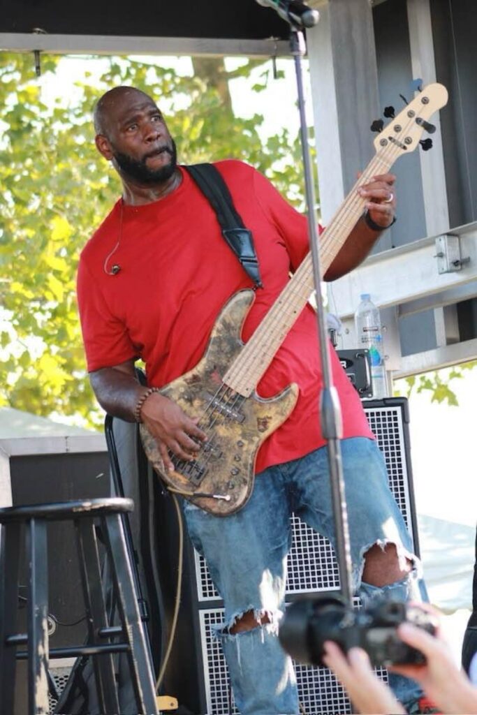 Jay Bratten playing a natural-finish bass guitar on an outdoor stage, wearing a red t-shirt and ripped jeans, with a photographer in the foreground