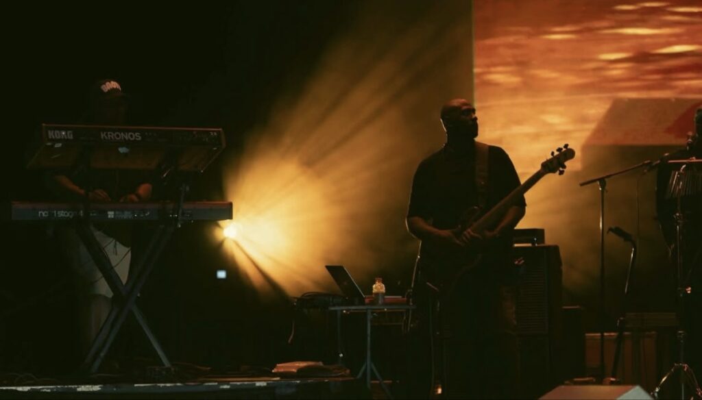Jay Bratten silhouetted playing bass on a dark stage lit by dramatic golden spotlights, with a keyboardist and Korg Kronos keyboards to the left