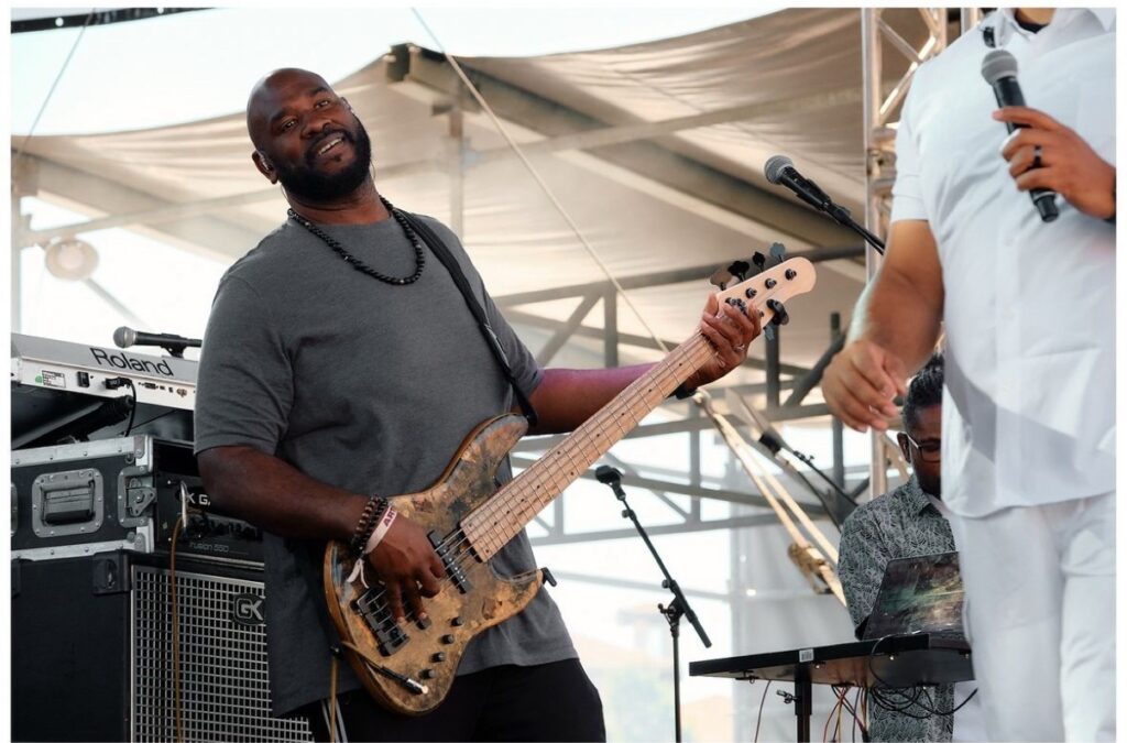 Jay Bratten smiling while playing a worn 5-string bass on an outdoor festival stage, with a Gallien-Krueger amp and Roland keyboard behind him