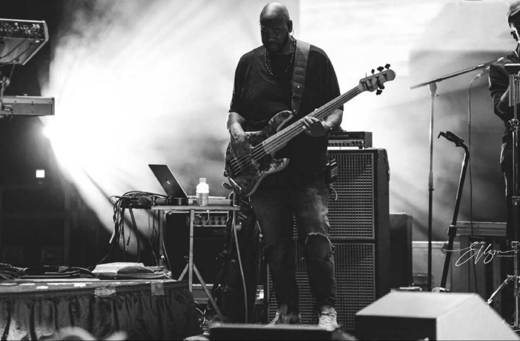Black and white photo of Jay Bratten playing bass center stage under bright backlights, wearing a beaded necklace and ripped jeans, with amp cabinets and a laptop stand nearby