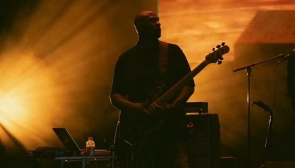 Jay Bratten silhouetted against intense orange stage lights while playing bass, with a laptop, water bottle, and amp cabinet on stage