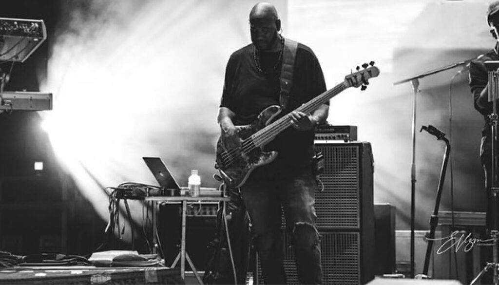 Black and white photo of Jay Bratten playing bass on a smoky stage with dramatic backlighting, laptop and amp cabinet beside him