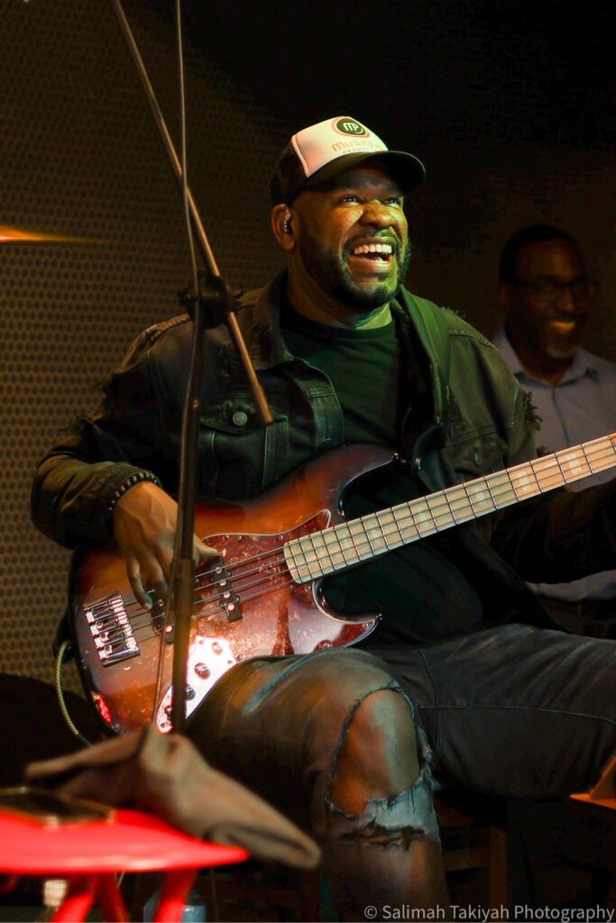 Jay Bratten smiling while playing a sunburst 5-string bass on stage, wearing a Musiqboy trucker cap and black denim jacket, with a fellow musician smiling behind him